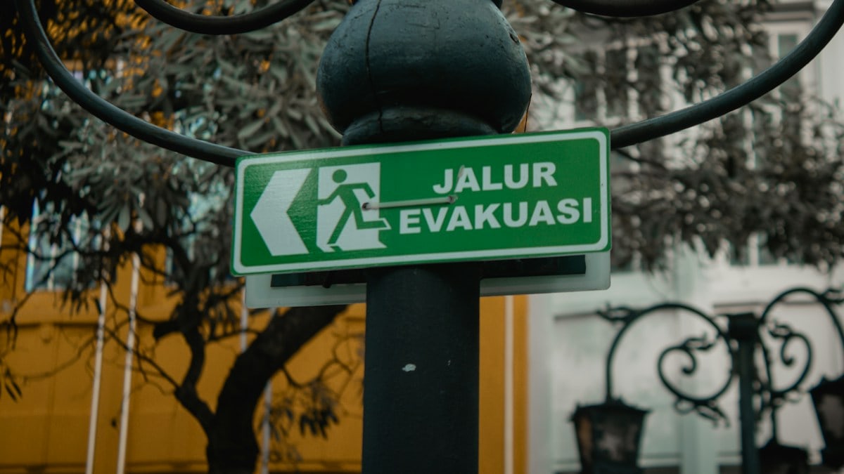 A bright green rectangular sign reading "JALUR EVAKUASI" (Evacuation Route) hangs from a weathered black metal pole adorned with intricate curved brackets. The sign features a bold white pictogram of a running figure, directing attention to the left. Set against a softly blurred backdrop of leafy branches and a yellow-walled building with ornate iron railings, the image balances functional urban infrastructure with subtle artistic detail, emphasizing clarity and safety in public spaces.